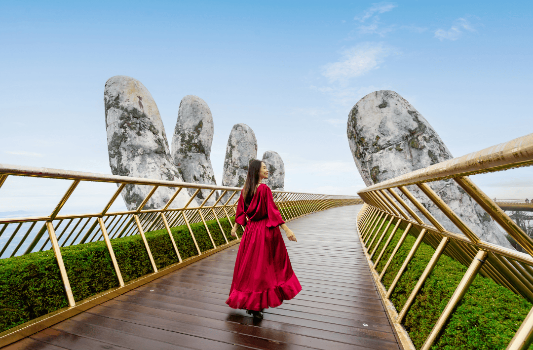 A woman in a flowing red dress walking on the Golden Bridge in Da Nang, Vietnam, with the iconic giant hands supporting the golden structure in the background.