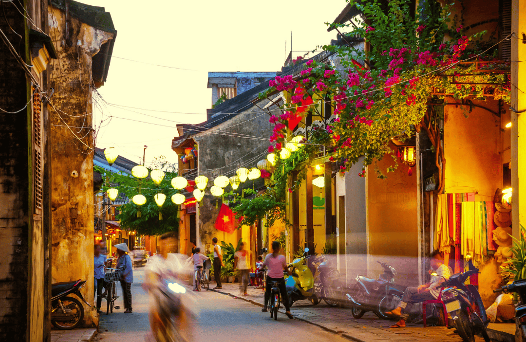  A charming street in Hoi An, Vietnam, lined with old houses, glowing lanterns, and bougainvillea flowers, with locals and tourists moving through the vibrant evening atmosphere.