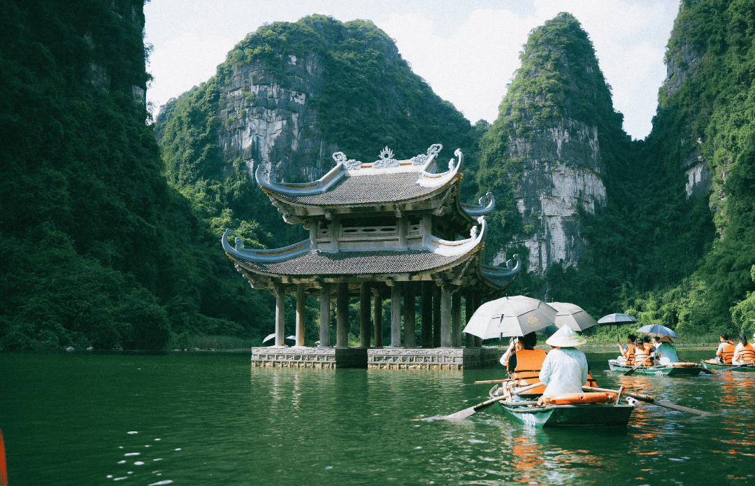 A serene scene of tourists rowing boats towards a traditional Vietnamese temple on stilts, surrounded by lush green limestone mountains in Ninh Binh, Vietnam.